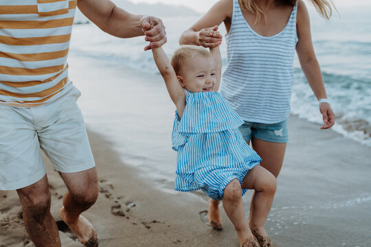 Parents holding hands with little toddler girl, walking on beach during beach holiday.