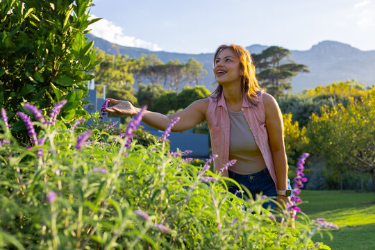 Adult African woman reaching for purple blooms with watch in park near blue-gray fence, copy space