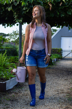 Woman walking gravel path in tended garden wearing shorts and boots holding pink watering can