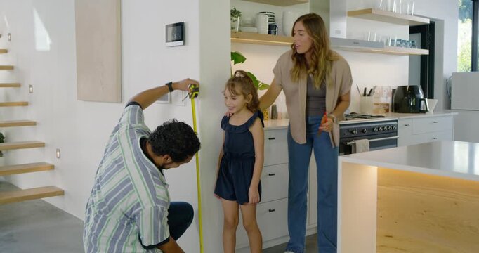 Diverse family measuring daughter in kitchen, dad with yellow tape, mom using pen to record height