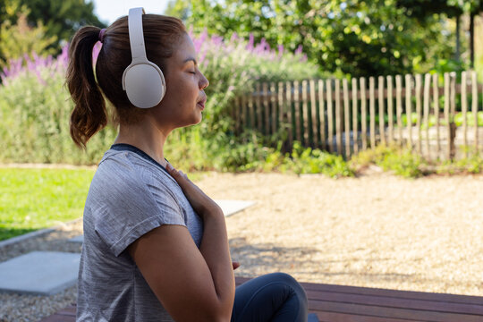 Woman sitting on wooden bench in garden in sportswear practicing breathing with headphones