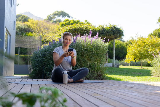 African American woman sitting on deck in garden with headphones, smartphone and bottle nearby