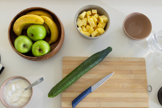 Bamboo board featuring cucumber and blue paring knife resting on light tabletop with fruit bowls