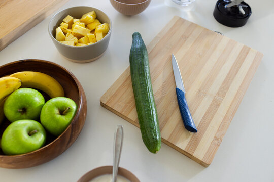 Long green cucumber lying on wooden cutting board on white counter, showing blue-handled knife
