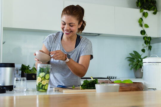 Adult woman smiling while pouring greens into blender cup at home kitchen with cucumbers and banana