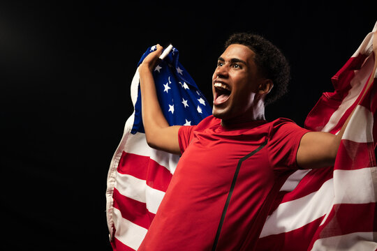 African American man wearing red athletic shirt holding American flag, shouting in dark studio