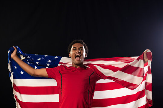 Adult African American male standing studio-lit holding US flag, shouting in red athletic shirt