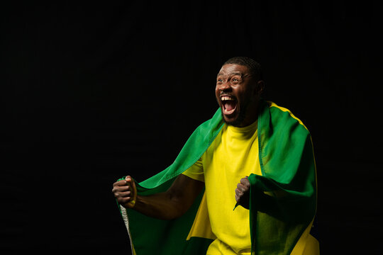 African man shouting clenching fists pulling green-yellow flag in yellow tee in studio, copy space