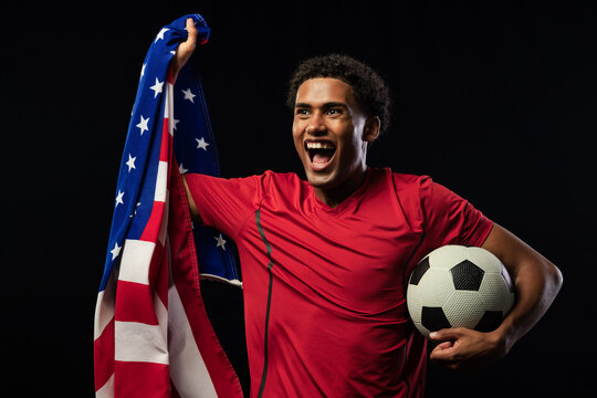 African American man wearing red jersey holding soccer ball draping US flag shouting in studio
