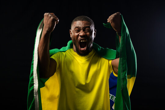 African man shouting, raising fists and gripping Brazilian flag while wearing yellow tee in studio
