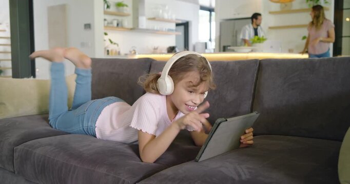 African-American child in headset lying on sofa tapping tablet giggling as parents prepping greens