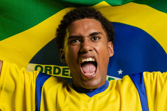 Male fan shouting, holding Brazilian flag behind shoulders in studio, wearing yellow jersey
