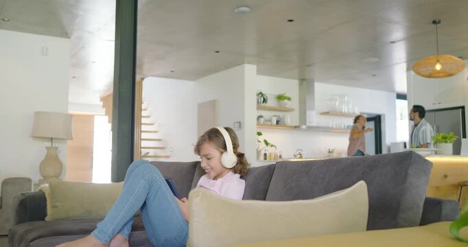 Diverse family child girl sitting on sofa at home with headset tapping device while parents moving