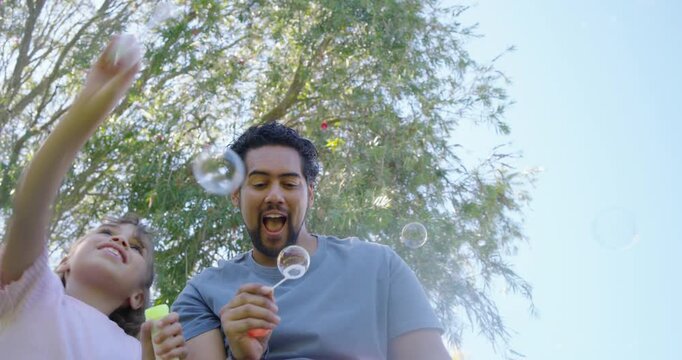 African American family playing while man dipping orange bottle, blowing bubbles under trees
