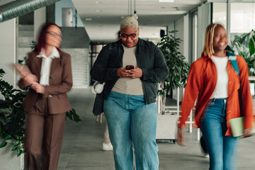Obraz premium Diverse businesswomen walking through a modern office building hallway, working with smartphones and interacting