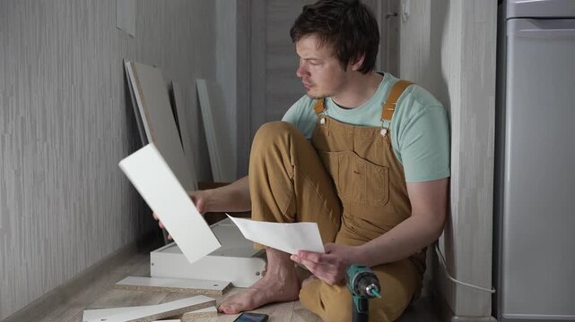 Frustrated man in overalls feeling stressed and confused while trying to assemble a new cabinet, carefully reading the difficult instructions and scratching his head in puzzlement