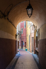Obraz premium A narrow vaulted passage opens to a quiet alley in Old Town Menton, France. Pastel facades, a wrought iron lantern, shutters, a small balcony, and a blue bucket stand out.