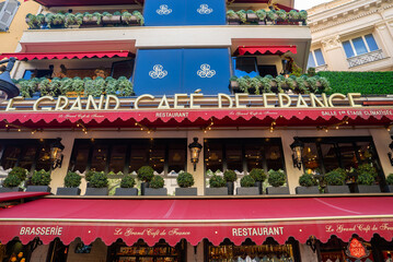 Obraz premium Street level view of Le Grand Cafe de France in Nice, France, with deep red awnings, navy balcony panels, string lights, and layered terraces in early evening.