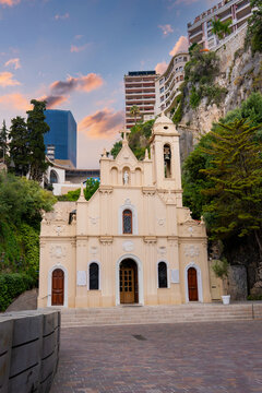 Chapelle Sainte Devote stands by the rocky cliff in Monaco at dusk, near the Sainte Devote curve, with arched doors and statues, framed by modern high rises.