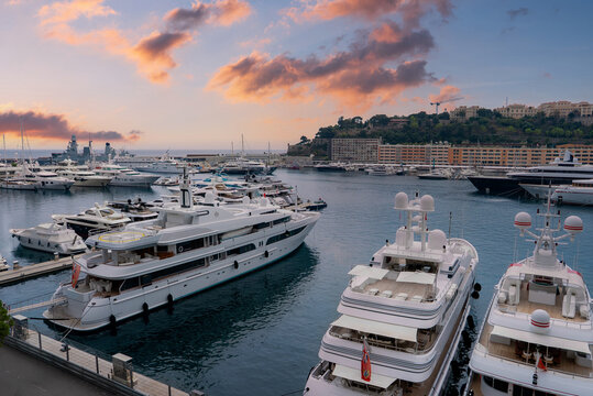 Port Hercule in Monaco shows superyachts, calm water, and cranes. Multi tiered apartments rise on a steep hillside as warm late day light reflects across the harbor.