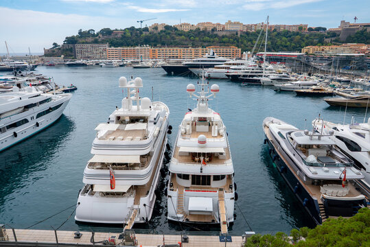 Luxury yachts line Port Hercule in Monaco, with Le Rocher and the Prince's Palace across the water. Daylight scene shows glossy decks, masts, cranes, and calm weather.