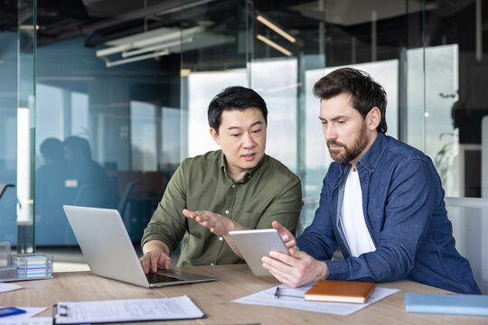 Two diverse businessmen are collaborating on a digital project at a modern office desk, one typing on a laptop and gesturing while discussing content displayed on a tablet held by his colleague
