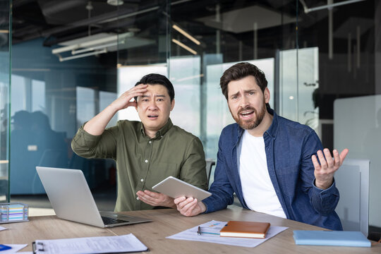 Diverse business partners sitting at an office desk, looking shocked and frustrated at the camera while experiencing confusion and stress about a mistake or problem