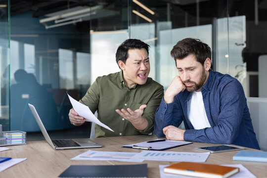 Upset asian executive manager berating a sad caucasian employee during a tense office meeting, focusing on themes of workplace conflict, stress, and harassment