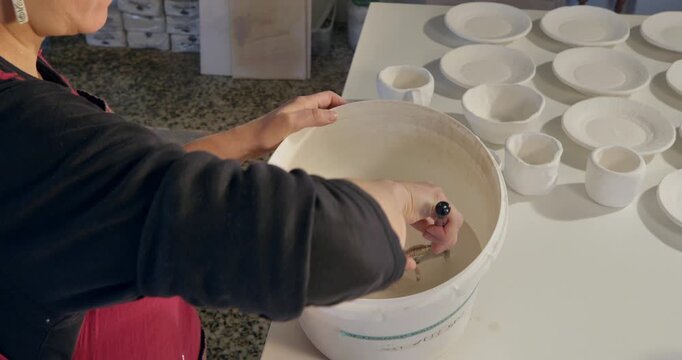 Crafting Pottery: A dedicated artisan stirs a bucket of liquid clay in a pottery workshop, with finished ceramic tableware elegantly arranged in the background.