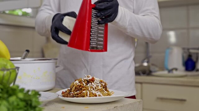 Close-up shot of hands in black gloves grating fresh cheese onto a plate of delicious homemade spaghetti. The person prepares a savory meal in a modern kitchen setting.