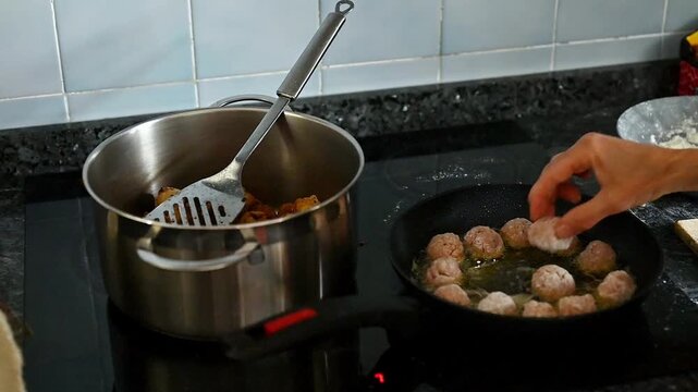 Home cooking process with meatballs frying in olive oil pan and pot in background, female hands adding raw meatballs, traditional Mediterranean kitchen recipe preparation, active food workflow