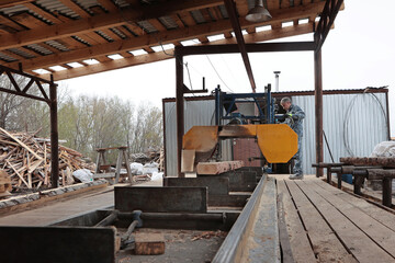 Fototapeta premium A worker controls a log sawing machine. Production of wooden pallets for heavy cargo transportation. An open-air locksmith workshop. The concept of woodworking.