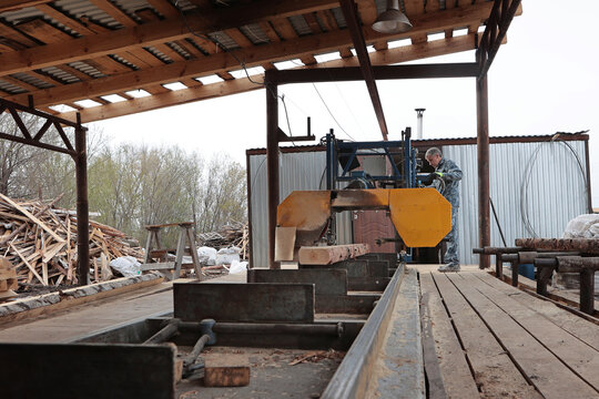 A worker controls a log sawing machine. Production of wooden pallets for heavy cargo transportation. An open-air locksmith workshop. The concept of woodworking.
