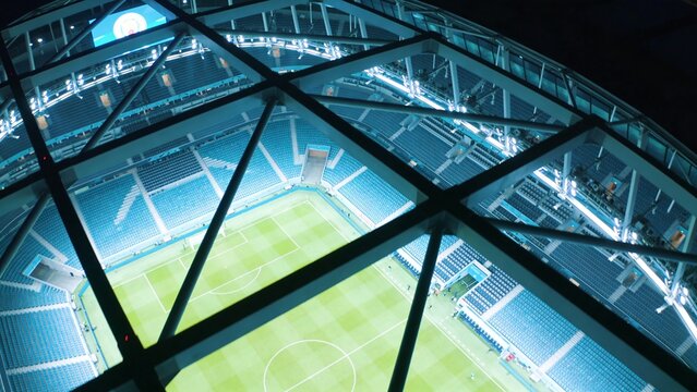 Framing high aerial view showing illuminated soccer pitch with goals, lights, roof truss at night