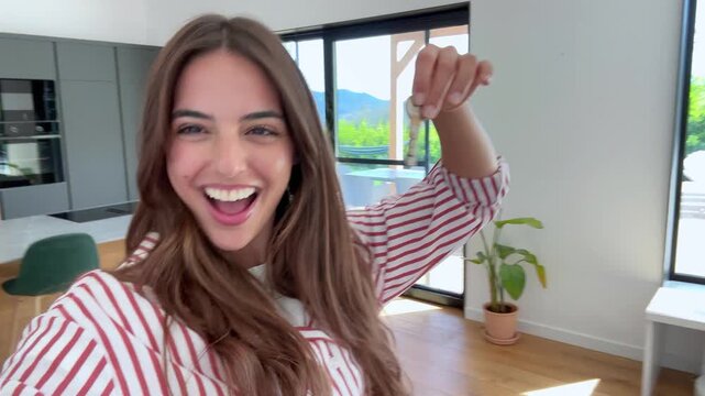 Excited young woman taking a selfie video while joyfully showing the keys to her new modern home, celebrating her first property purchase and the beginning of a new chapter in life