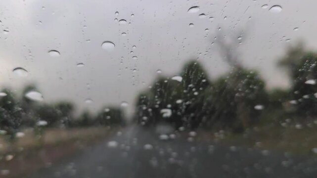Raindrops falling on a car windshield with a blurred view of a rural road and forest under a gloomy sky 120fps 4K Slow Motion Captured in Cinematic High-Detail Clip