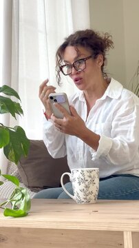 Cheerful mature woman with glasses sitting on the sofa at home while having an engaging video conference on her smartphone, smiling and gesturing with her hands in a bright living room