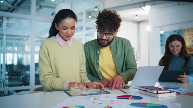 Business partners analyzing digital report on tablet screen. African American man checking charts and pointing at statistics near desk. Female colleague working on laptop in background.