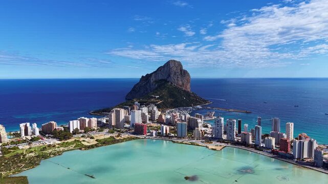 Aerial view of Calpe, Spain showing the iconic Penon de Ifach rock formation, turquoise Mediterranean Sea, salt lagoon, and modern resort buildings on the Costa Blanca