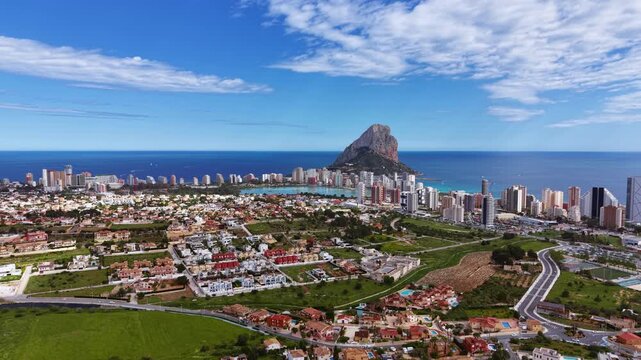 Panoramic view of Calpe, Spain, showcasing the iconic Penon de Ifach rock formation, coastal skyline, residential neighborhoods, and turquoise Mediterranean Sea under blue sky