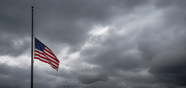 American flag at half mast against dark stormy clouds representing national mourning and sadness