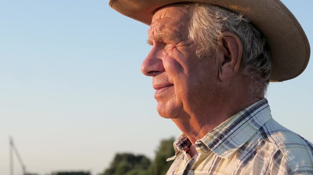 Close up old farmer with gray hair wearing cowboy hat and plaid shirt. Man gazing into the distance as the sun sets, with a slight smile and wrinkles illuminated by warm light.