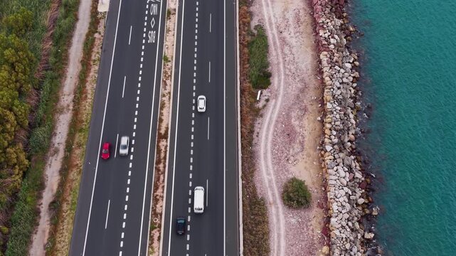 Top-down view of a multi-lane coastal highway with cars driving alongside a rocky breakwater and vivid turquoise sea, showing road markings and gantry signs