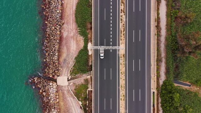 Overhead view of a two-lane coastal road with cars driving alongside a rocky breakwater, turquoise sea water, sandy beach, and green vegetation