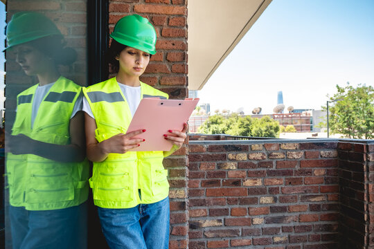 Young female safety inspector wearing green safety helmet and yellow safety vest, holding pink folder while supervising construction site safety and giving instructions from balcony