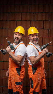 Happy middle aged male construction brothers twins workers in orange overalls and yellow helmets holding drills. Professional builders posing in front of brick wall during house renovation work
