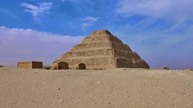 World's oldest pyramid, Step Pyramid of Djoser at Saqqara, Egypt, rising above ancient ruins and stone blocks with arched entrance wall under blue sky.