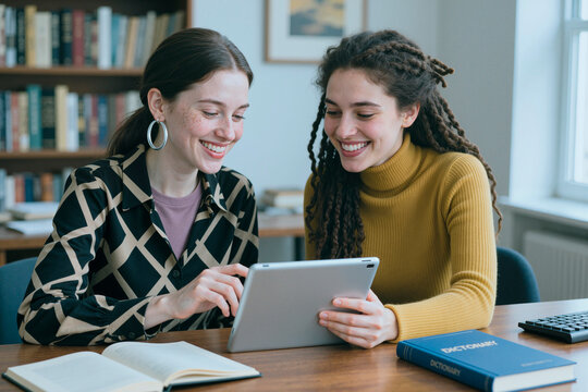 Young woman and young woman studying on tablet in library, collaborating on research and digital learning. Ideal for education technology, tutoring, academic content
