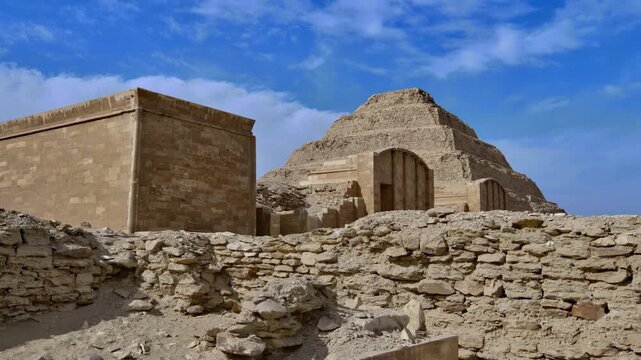 Step Pyramid of Saqqara, Egypt, designed by architect Imhotep for pharaoh Djoser, towering over archaeological ruins and stone debris under partly cloudy sky.