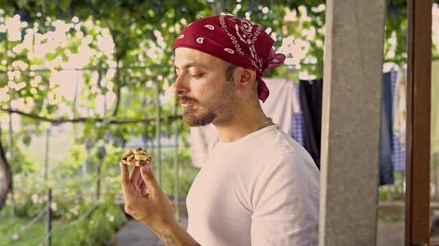 A young man wearing a red bandana and a white t-shirt savors a delicious sweet snack, possibly a homemade cookie or pastry, while enjoying the outdoors.